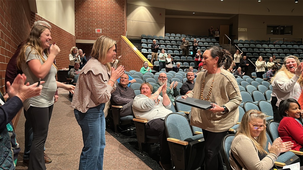 A teacher is surprised with an award as all around her clap and cheer. 