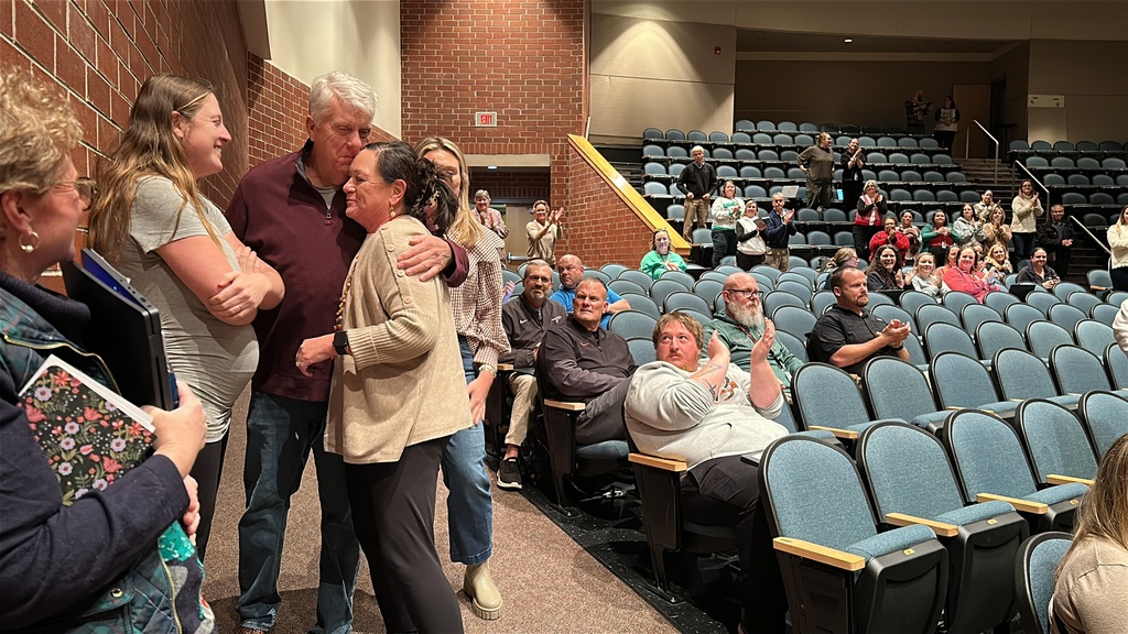A teacher is surprised with an award as all around her clap and cheer. 