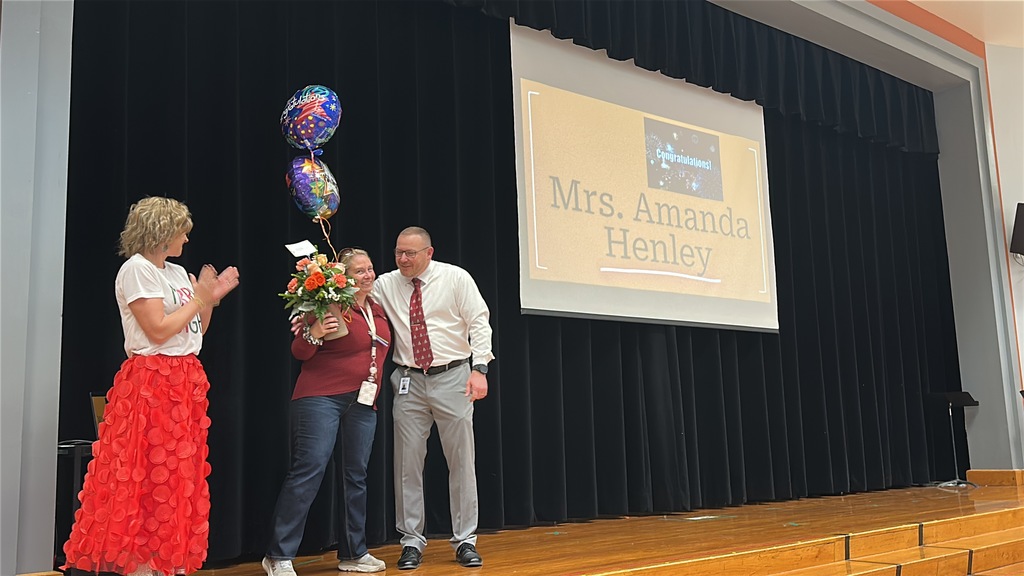A man and woman congratulate a woman holding flowers. A slide behind them reads COngratulations Mrs. Amanda Henley.