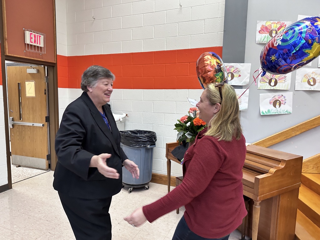 One woman reaches to hug another woman holding flowers. 