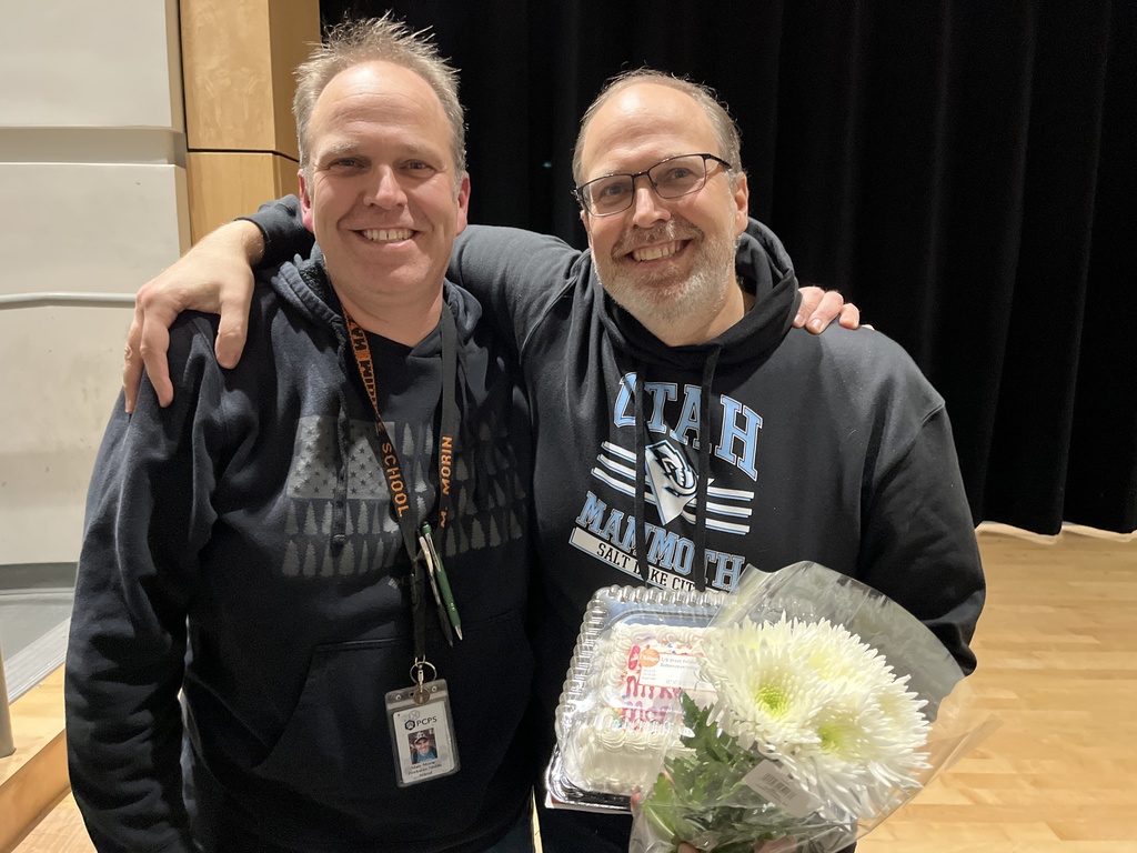 Two men pose for a photo.  One has flowers and a cake.