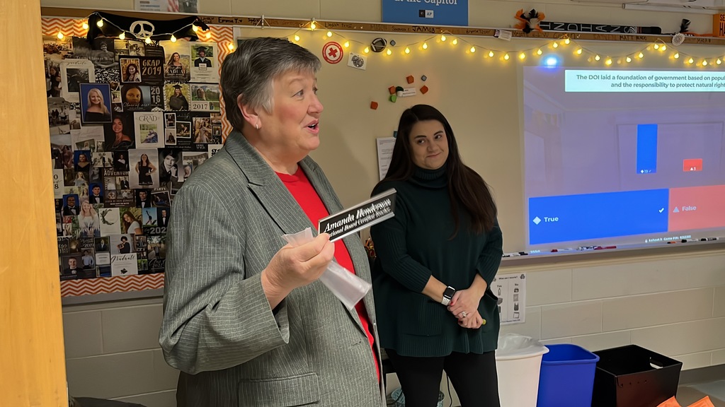 A woman presents an National Board Certified Teacher name plaque to an educator.