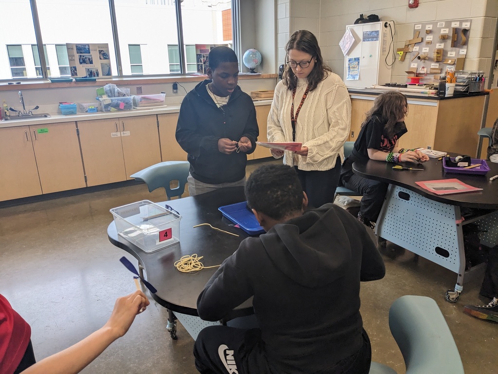 Students work on an assembly line making airplanes in class.  A teacher helps.