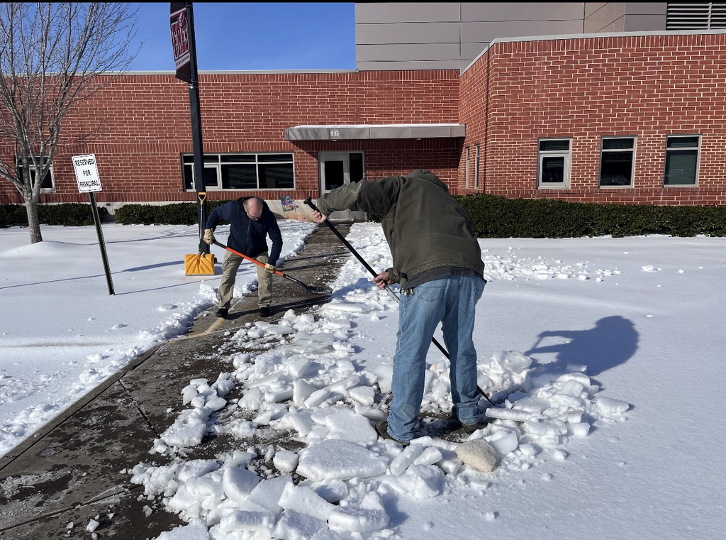 Two people clear a school sidewalk of snow and ice. 