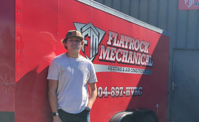 A student poses for a photo in front of a company logo representing where he is doing a Youth Registered Apprenticeship.