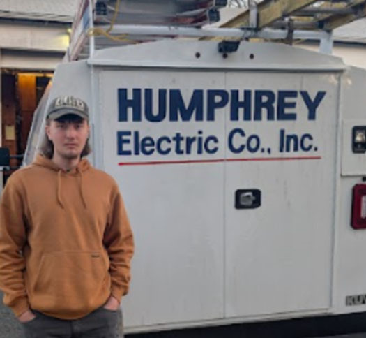 A student poses for a photo in front of a company logo representing where he is doing a Youth Registered Apprenticeship.