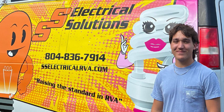 A student poses for a photo in front of a company logo representing where he is doing a Youth Registered Apprenticeship.