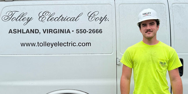 A student poses for a photo in front of a company logo representing where he is doing a Youth Registered Apprenticeship.