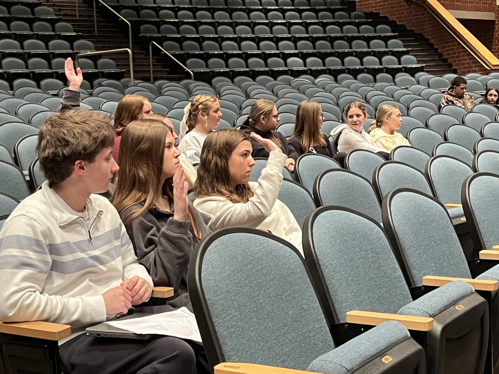 Students sit in chairs in an auditorium. A few of them are raising a hand.