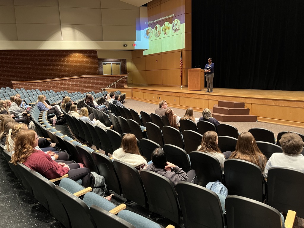 A woman onstage speaks to students sitting in an auditorium. 
