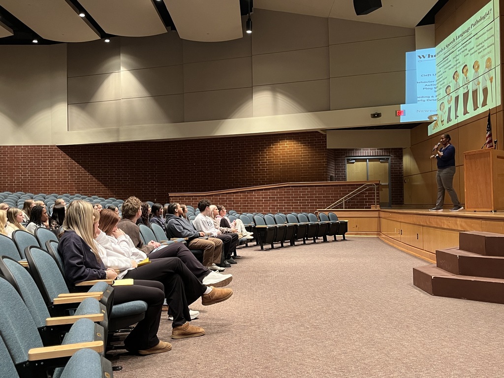 A woman onstage speaks to students sitting in an auditorium. 