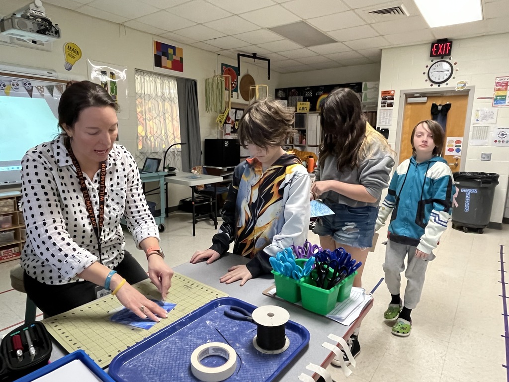 A teacher helps cut holes on a student's work so he can attach art pieces. Other students wait in line. 