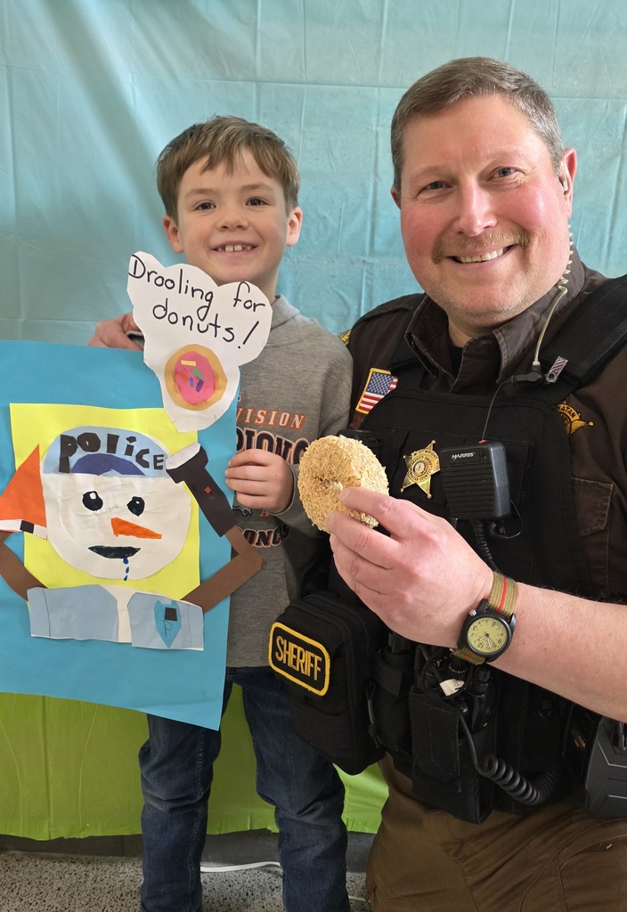 A deputy holding a donut poses for a photo with a child holding a policeman snowman art piece. 