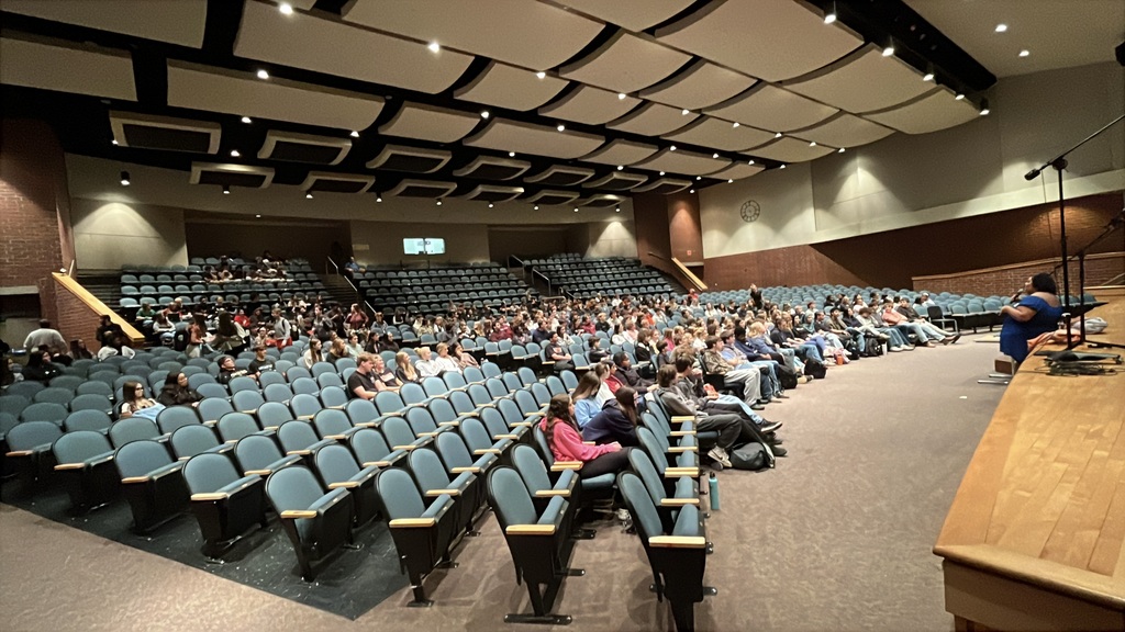 A guest presenter speaks to students in a crowded auditorium.