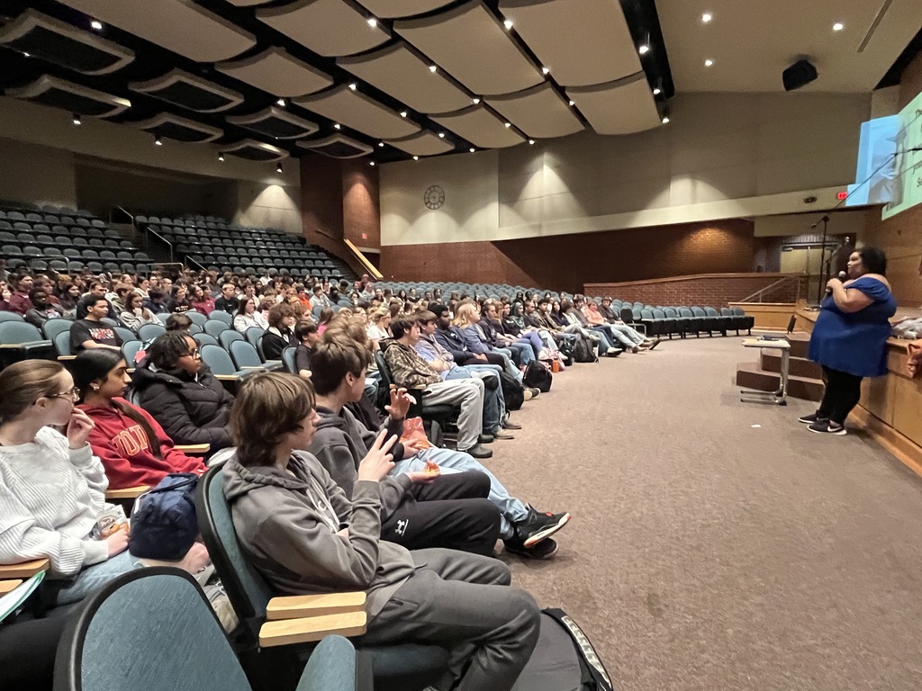 A guest presenter speaks to students in a crowded auditorium.