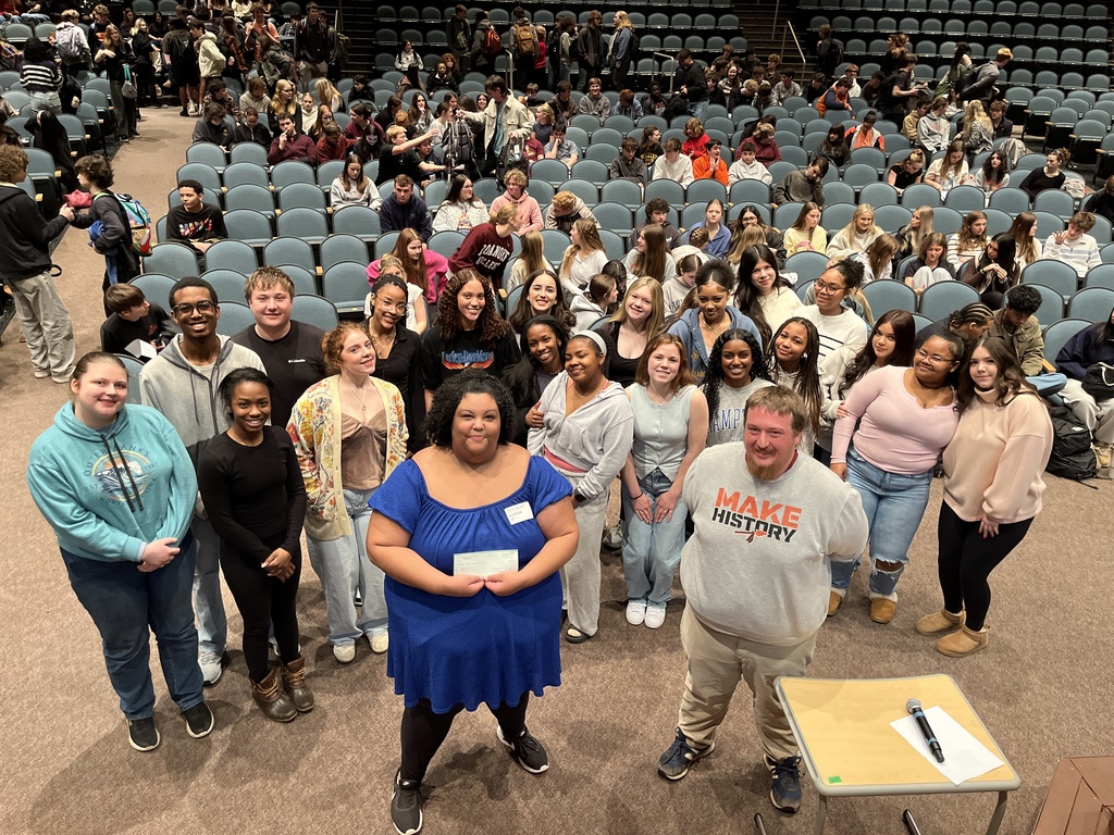 A guest presenter poses with a group of students in a crowded auditorium.