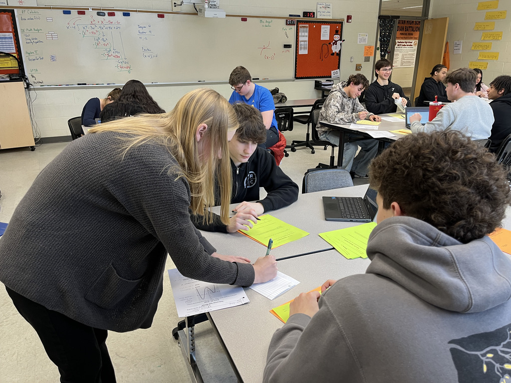Algebra II classes work in groups to review polynomial operations and division through rotating stations. A student teacher helps.