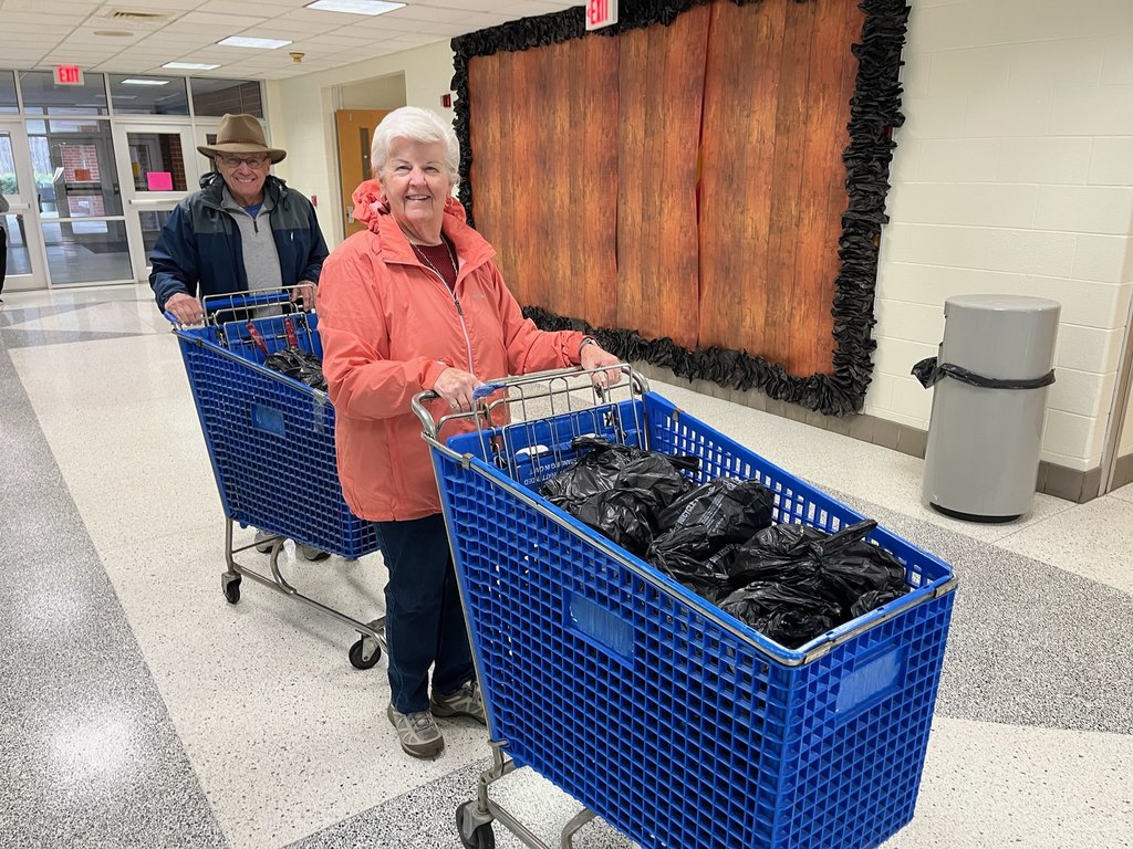 A man and woman in a school push grocery cards full of black bags full of food for children.