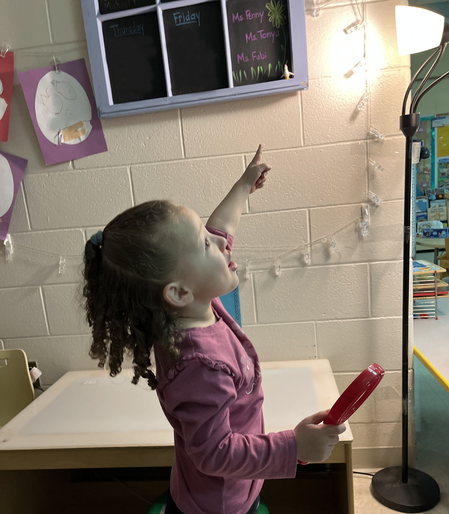 A preschooler  uses a large magnifying glass to help find a missing class toy.