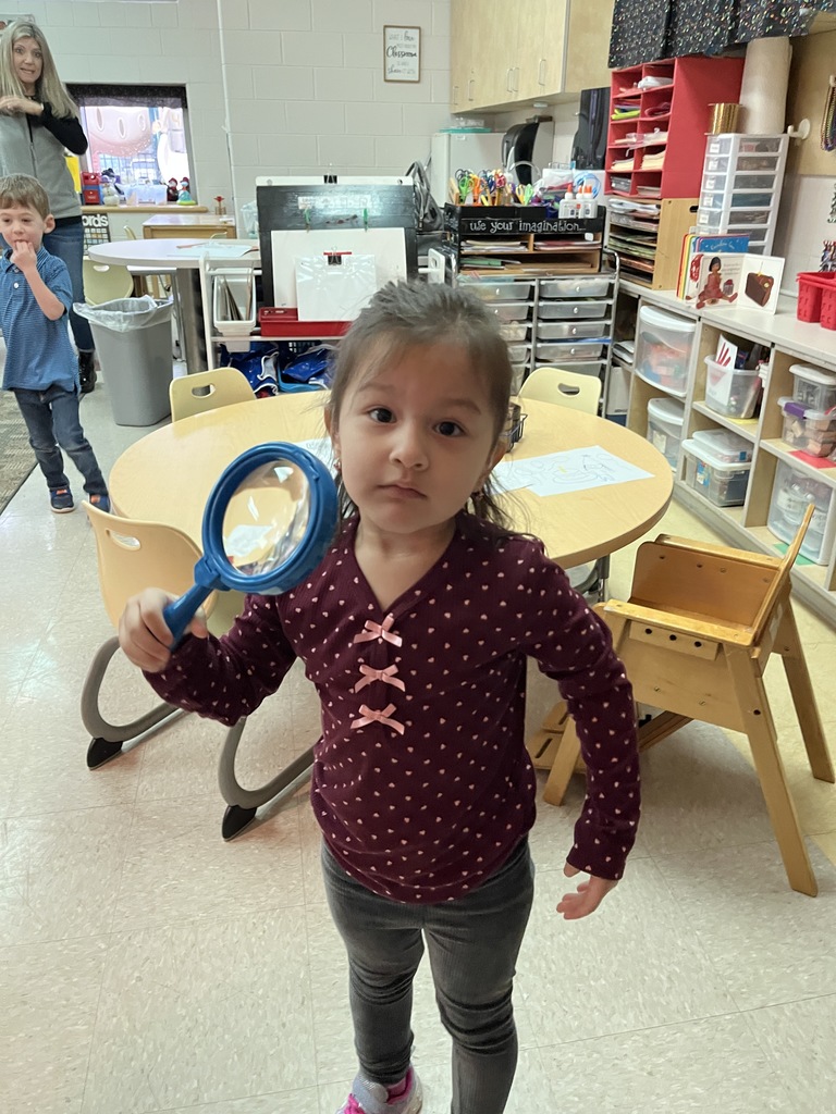 A preschooler  uses a large magnifying glass to help find a missing class toy.