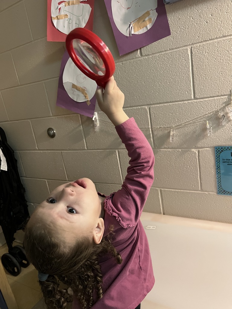 A preschooler  uses a large magnifying glass to help find a missing class toy.