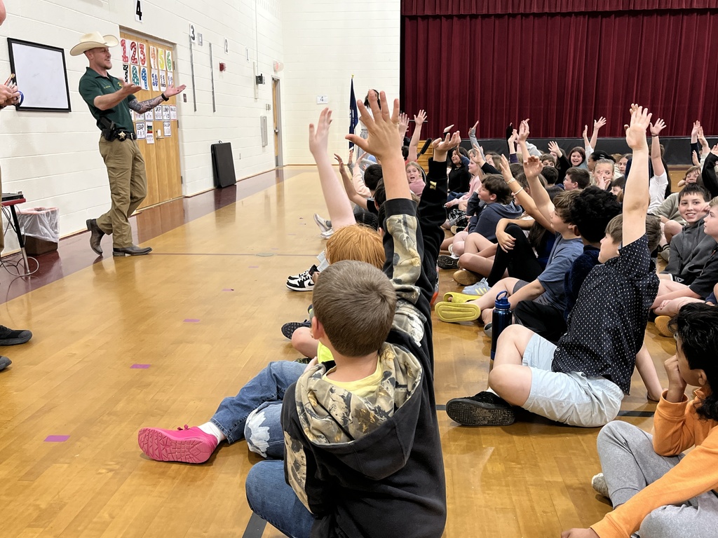 A detective speaks to a group of students about cybersecurity in a school assdembly. Many of the students are raising their hands.