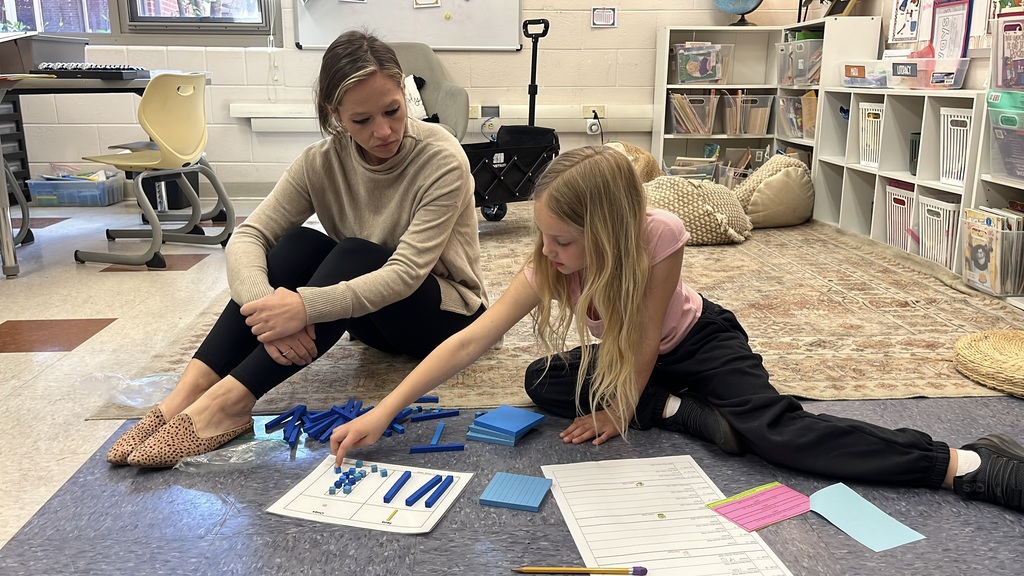 A child works on an egg-hunt themed math activity about place values. A teacher observes.