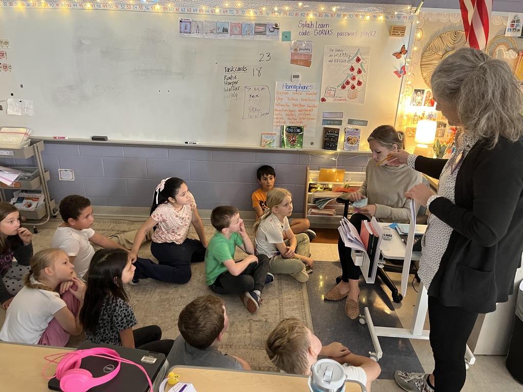 Two teachers speaking to a calss sitting on a carpet. 