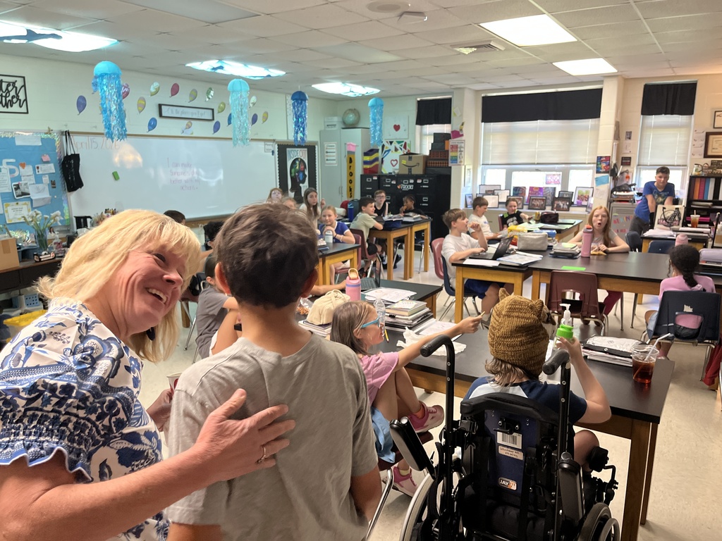 A teacher greets a student in a classroom full of other students.