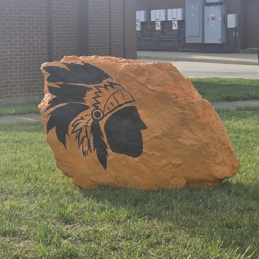 A large rock on a lawn has been painted orange with the silhouette of a Native American painted on it. 