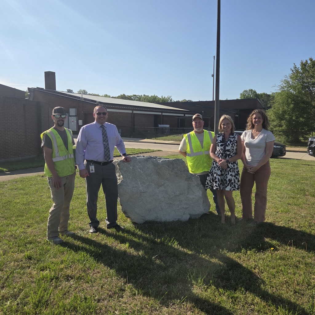 Five adults pose with a large rock in front of a school.