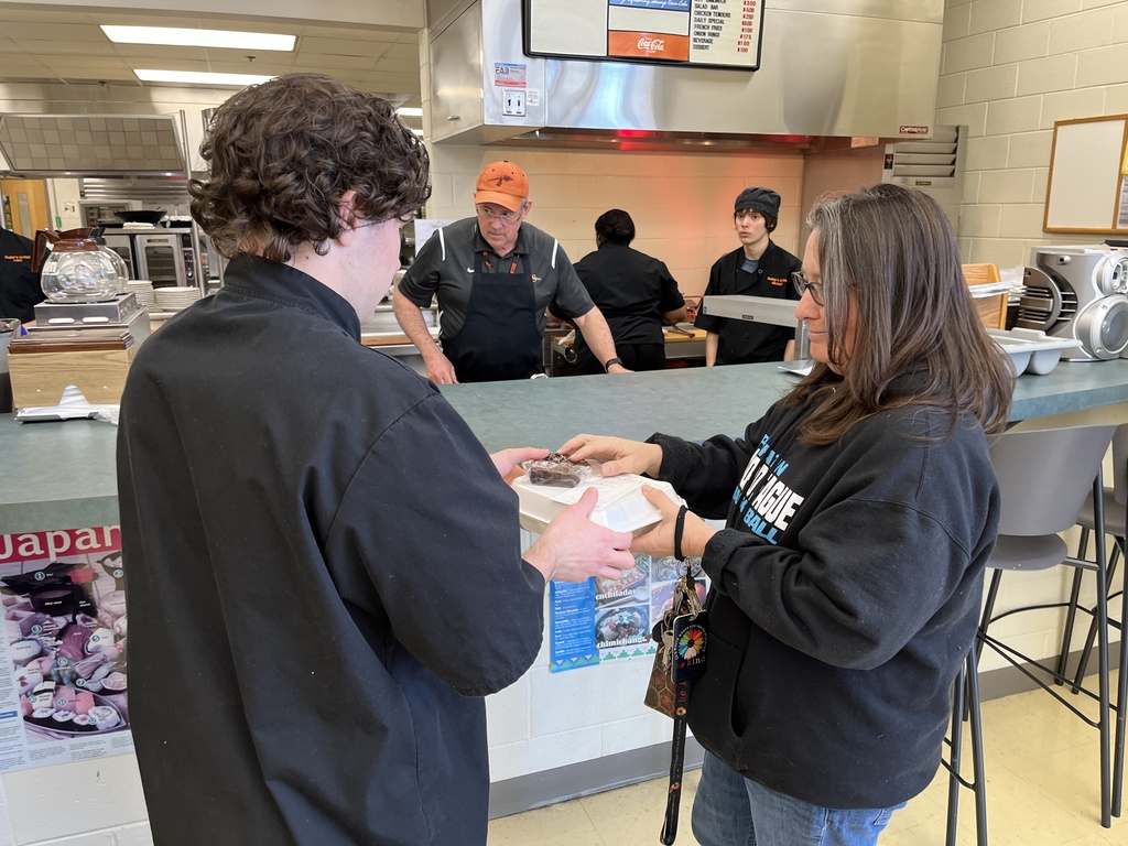 A teacher picks up her meal in a culinary cafe, from a student. A teacher and students work in the background.