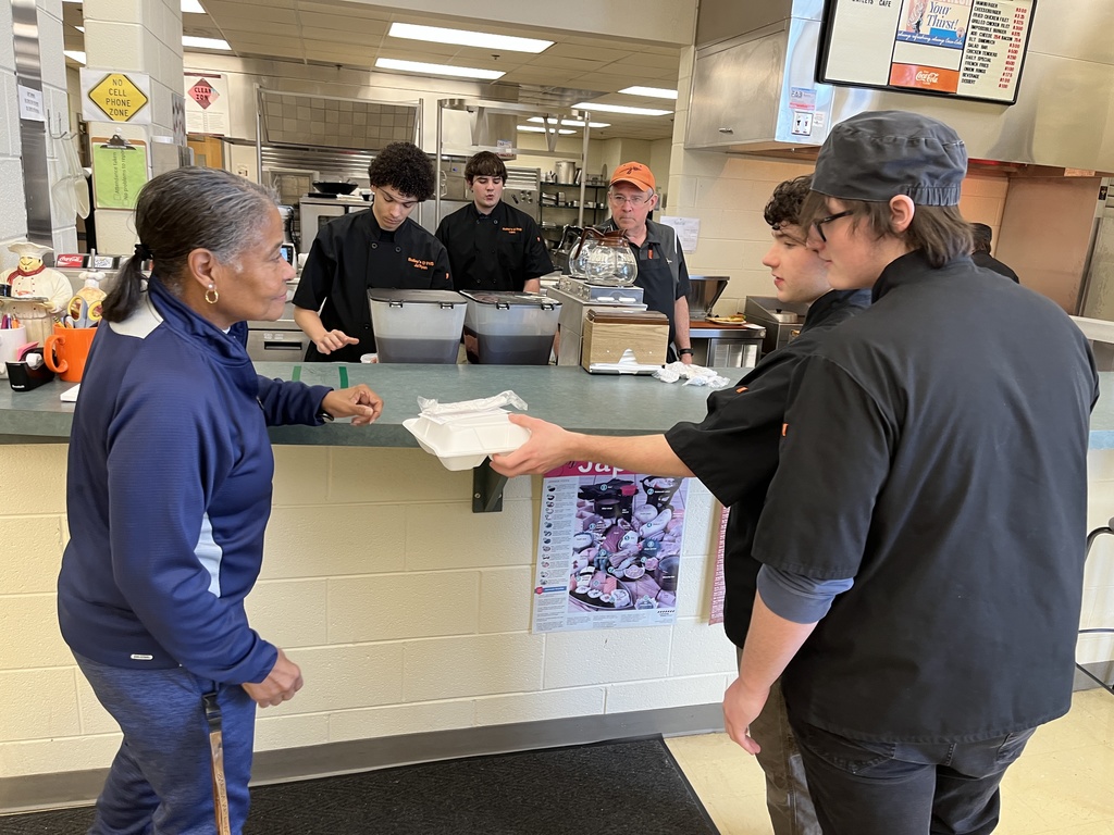 A teacher picks up her meal in a culinary cafe, from a student. A teacher and students work in the background.