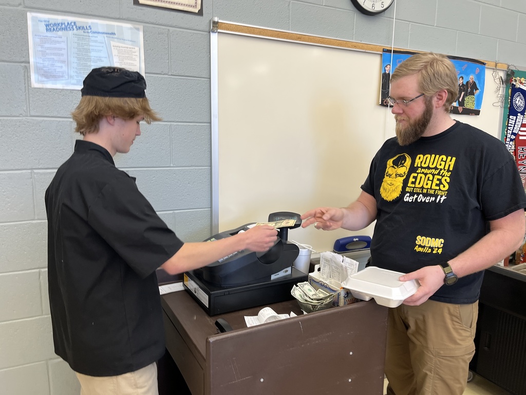 A teacher pays for his meal in a culinary cafe, giving the money to a student cashier.