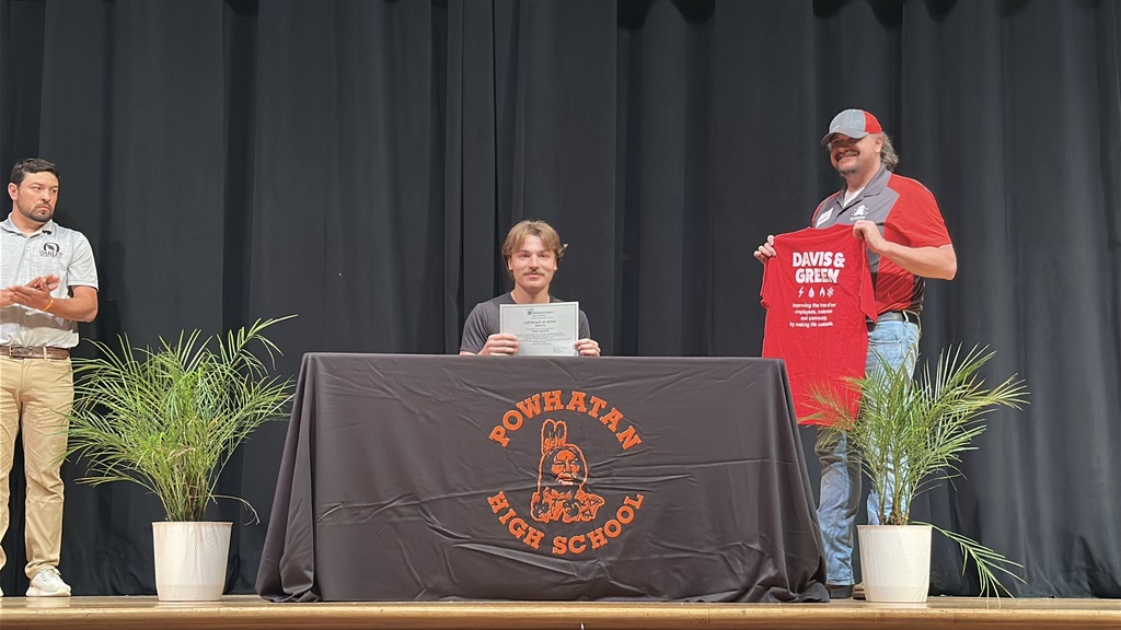 A  student holds up a piece of paper and poses  at a high school career and workforce development signing day. A man stands behind him.