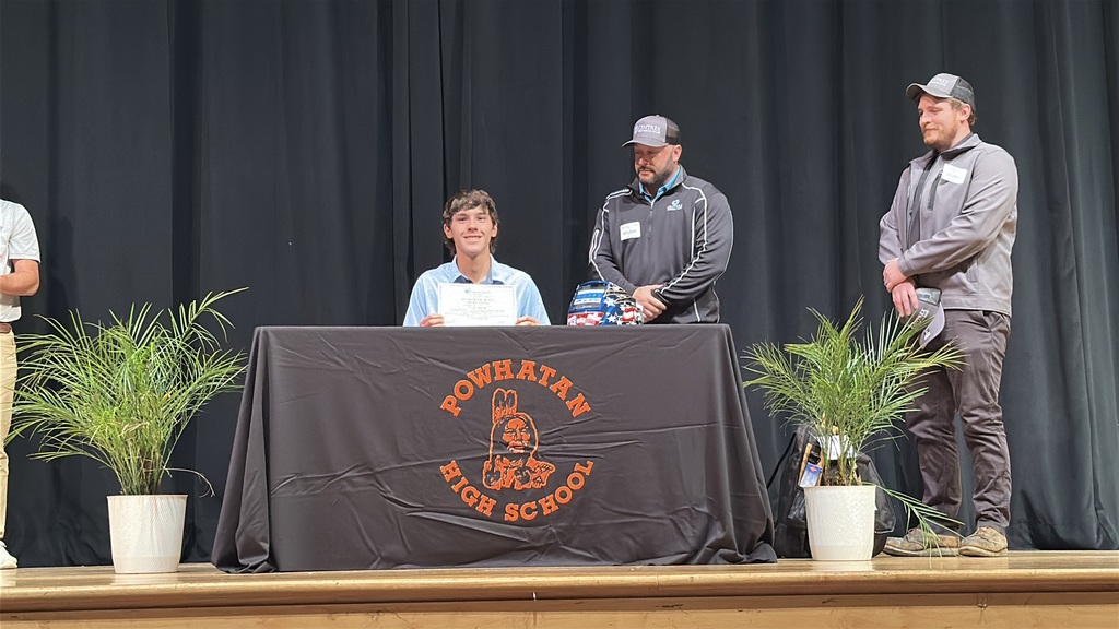 A  student holds up a piece of paper and poses  at a high school career and workforce development signing day. Two men behind him.
