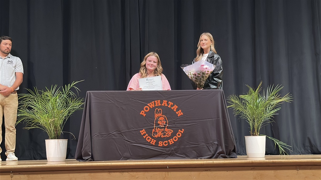 A  student holds up a piece of paper and poses  at a high school career and workforce development signing day. A woman stands behind her..