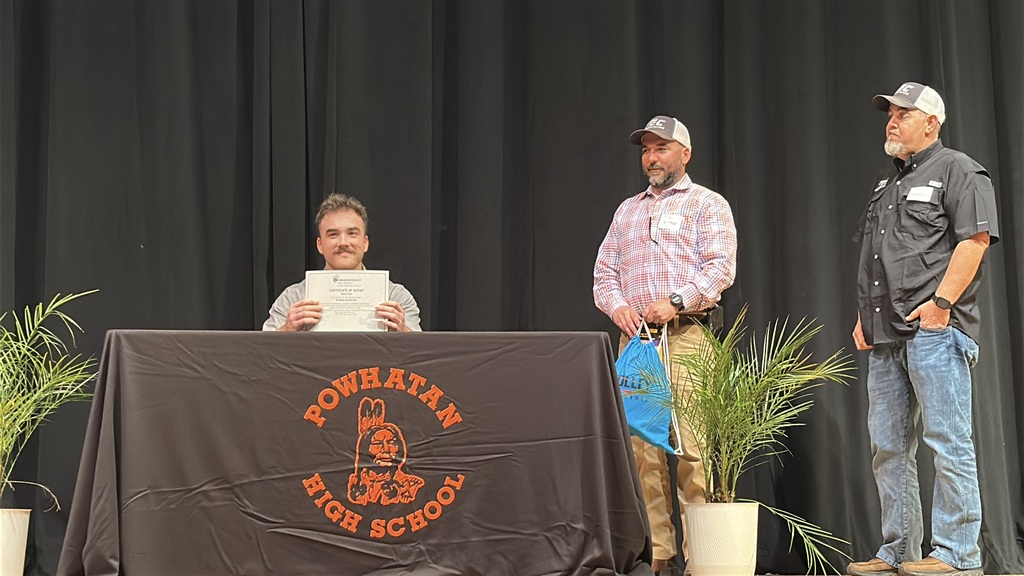 A  student holds up a piece of paper and poses  at a high school career and workforce development signing day. A man stands behind him.