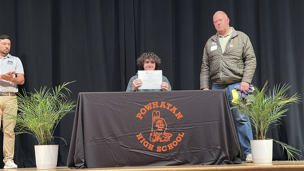 A  student holds up a piece of paper and poses  at a high school career and workforce development signing day. A man stands behind him.