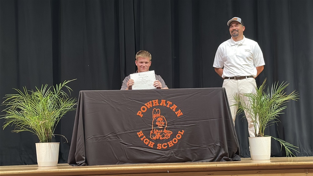 A  student holds up a piece of paper and poses  at a high school career and workforce development signing day. A man stands behind him.