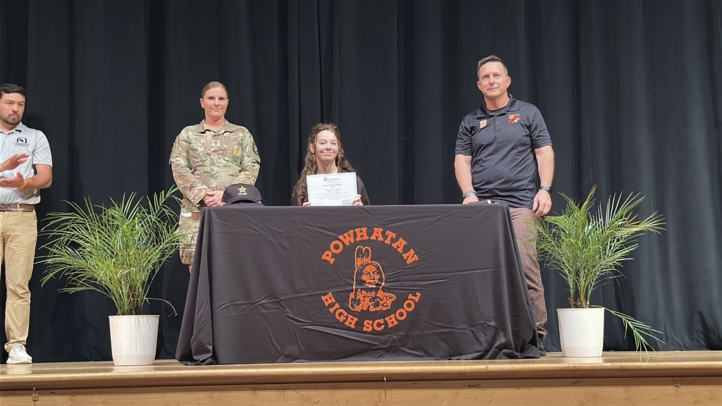 A  student holds up a piece of paper and poses  at a high school career and workforce development signing day. A man and a woman stand behind her.