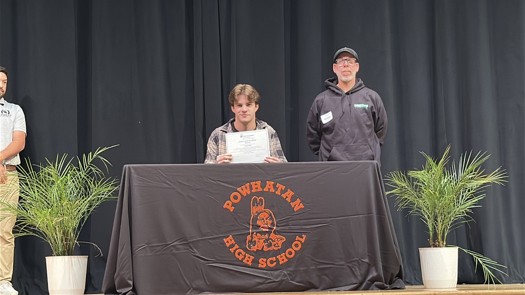 A  student holds up a piece of paper and poses  at a high school career and workforce development signing day. A man stands behind him.