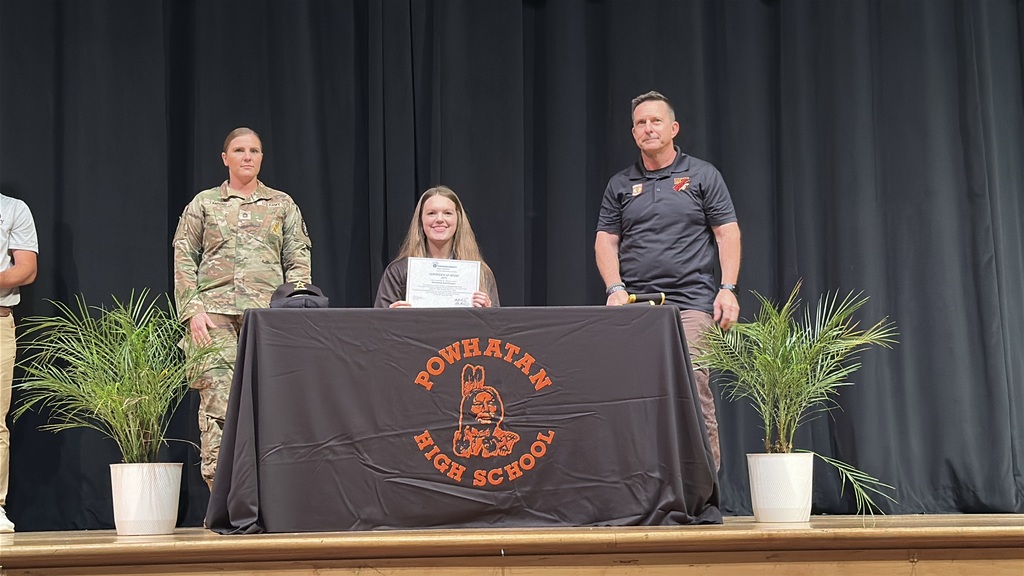 A  student holds up a piece of paper and poses  at a high school career and workforce development signing day. A man and a woman stand behind her.