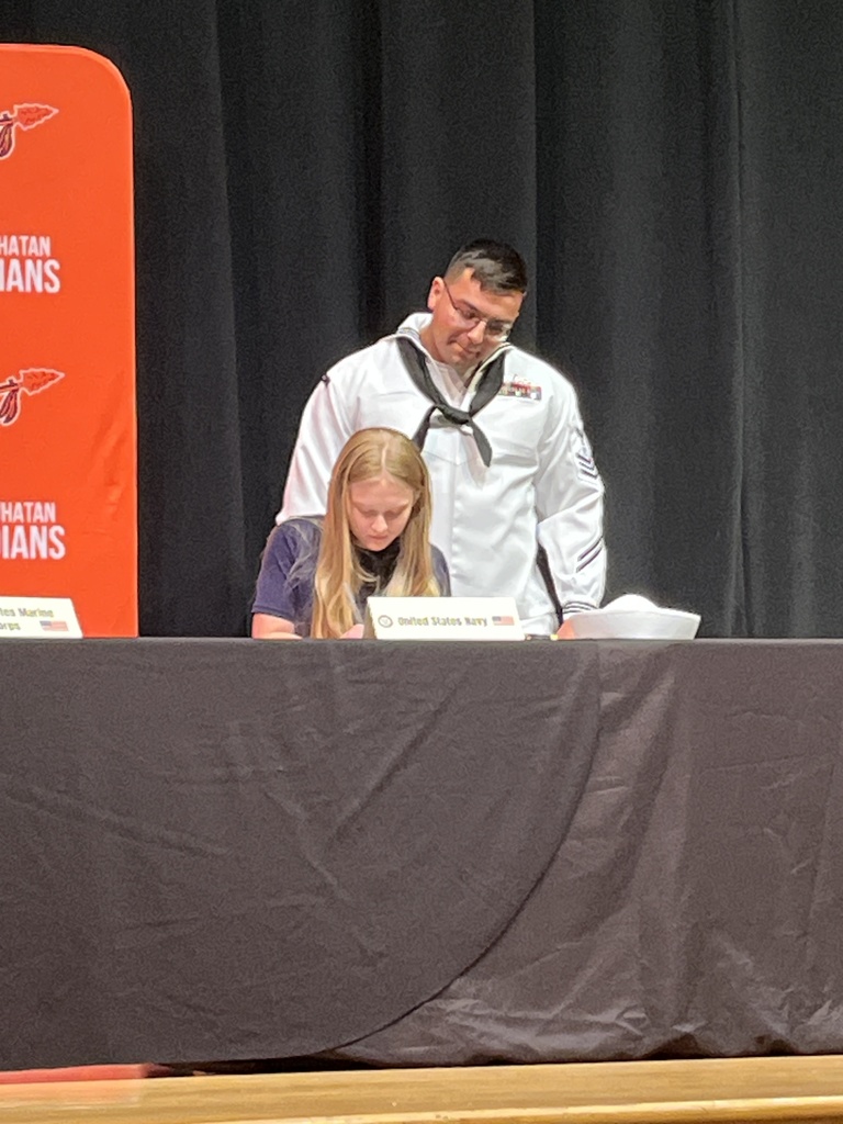 A military service member stands behind a student signing a letter of intent at a military signing day ceremony.