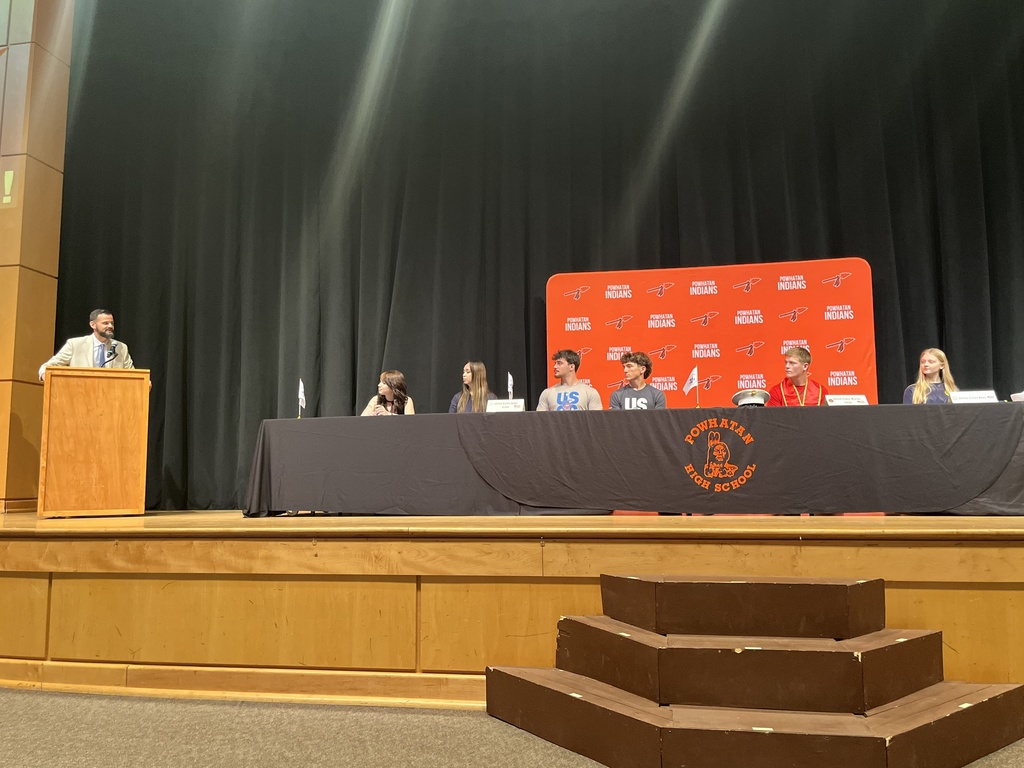 A man stands behind a podium and speaks to a group onstage at a military signing day ceremony.