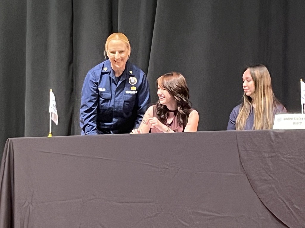 A military service member stands behind a student signing a letter of intent at a military signing day ceremony.