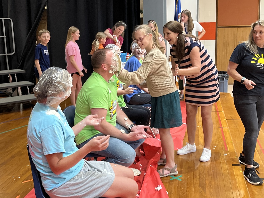 A group of adults sit in a line of chairs as part of a pie-in-the face event. A child pushes a pie into one of their faces. 