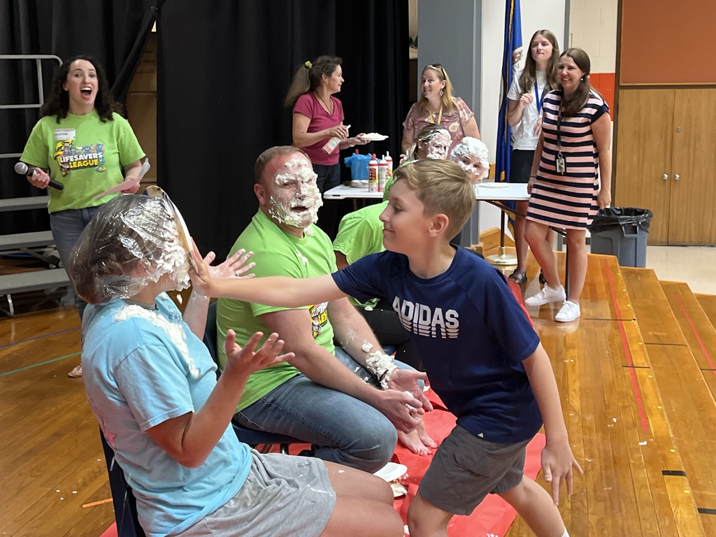 A group of adults sit in a line of chairs as part of a pie-in-the face event. A child pushes a pie into one of their faces. 