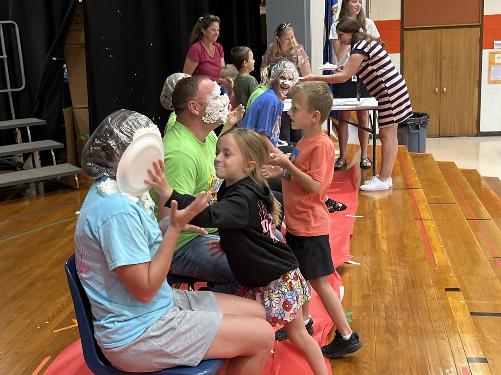 A group of adults sit in a line of chairs as part of a pie-in-the face event. A child pushes a pie into one of their faces. 