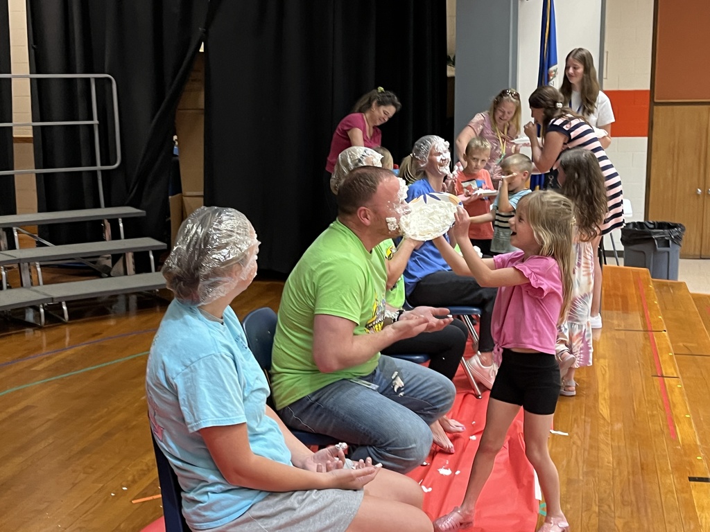 A group of adults sit in a line of chairs as part of a pie-in-the face event. A child pushes a pie into one of their faces. 